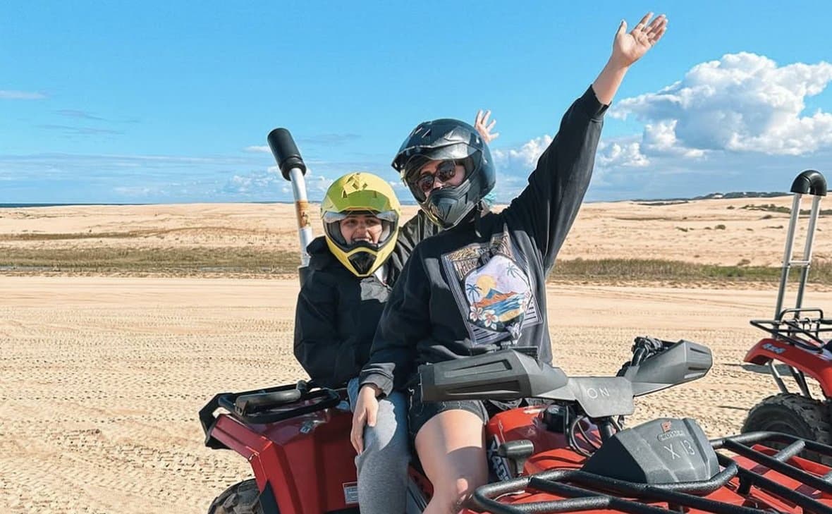 Two people on a quad bike on the sand dunes, arms in the air